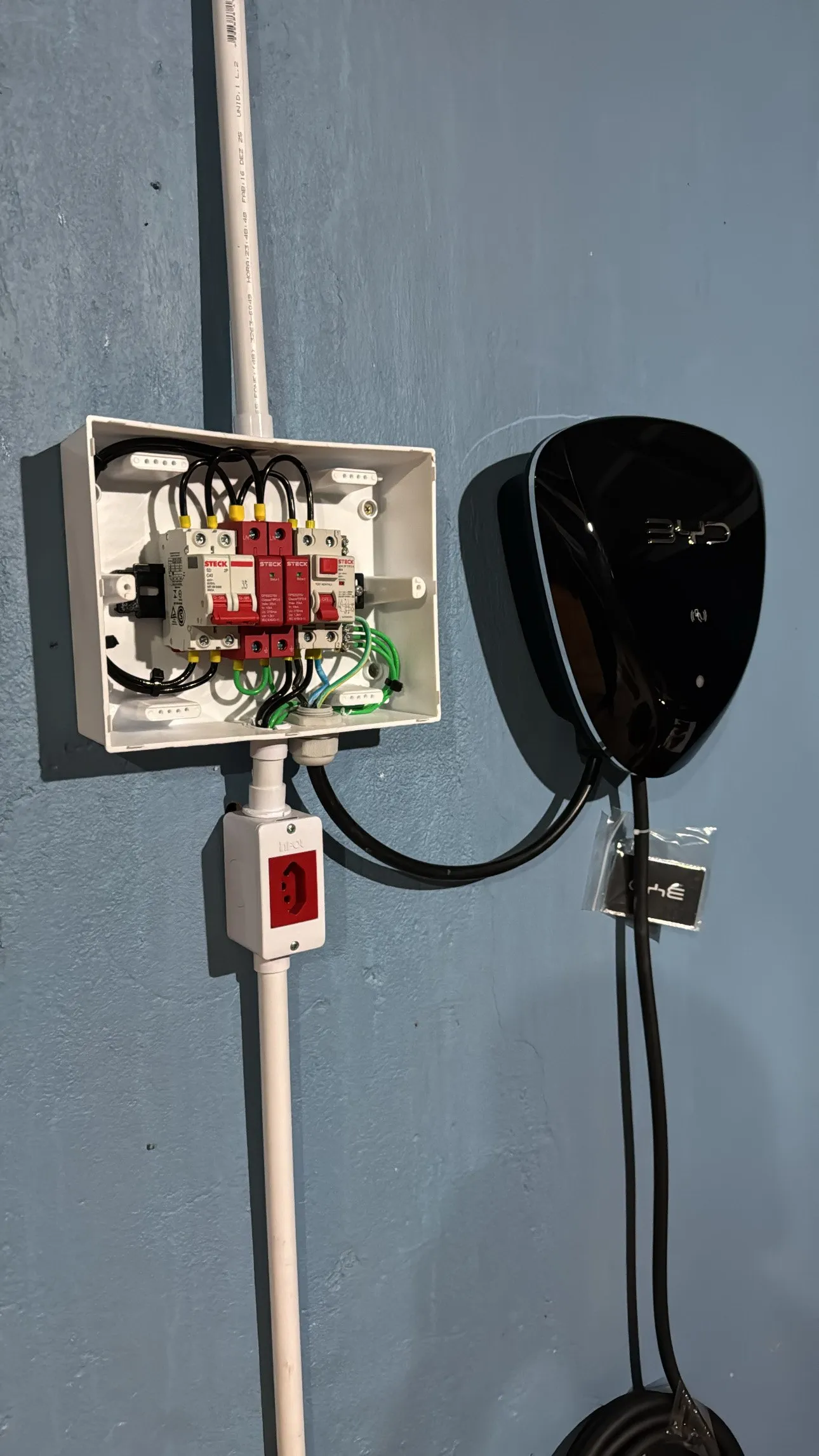 Wallbox preto instalado em parede azul com quadro de proteção e eletroduto aparente